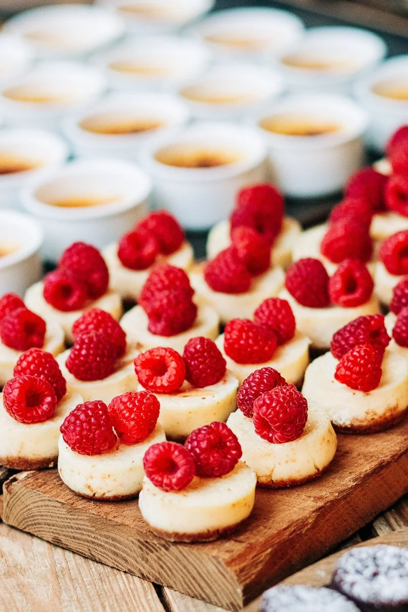 Mini cheesecakes topped with fresh raspberries are arranged on a wooden board. Blurred rows of crème brûlée in white ramekins are in the background.