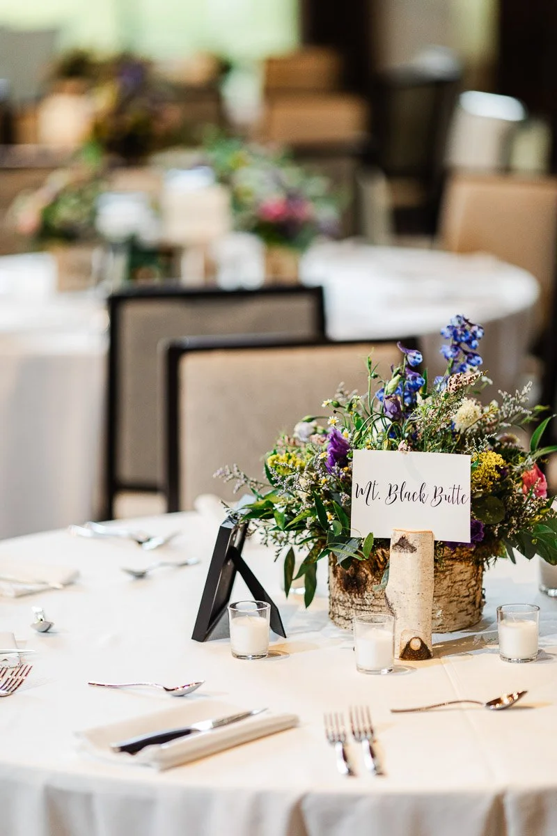 Elegant wedding table with a floral centerpiece featuring purple, yellow, and green blooms. A "Mt. Black Butte" sign is prominent, giving a rustic touch.