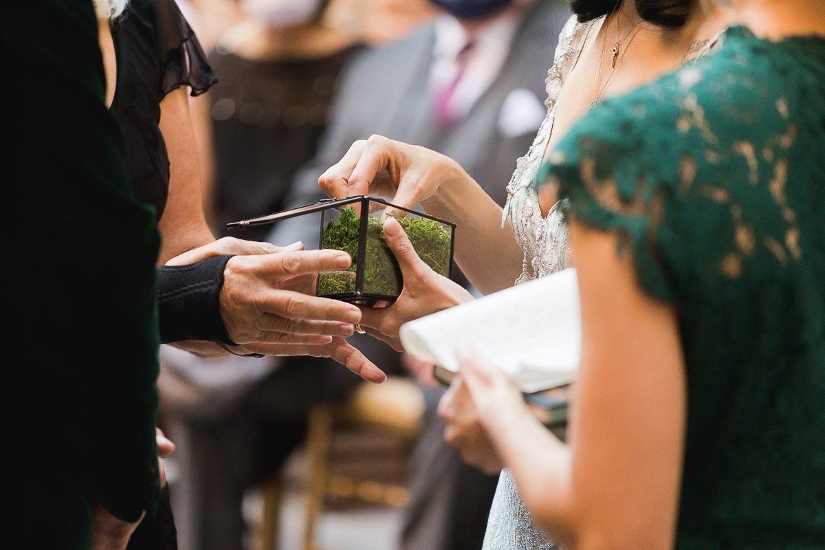 A close-up of hands at a wedding ceremony holding a small glass box with moss. The scene feels intimate and ceremonial, with formal attire visible.
