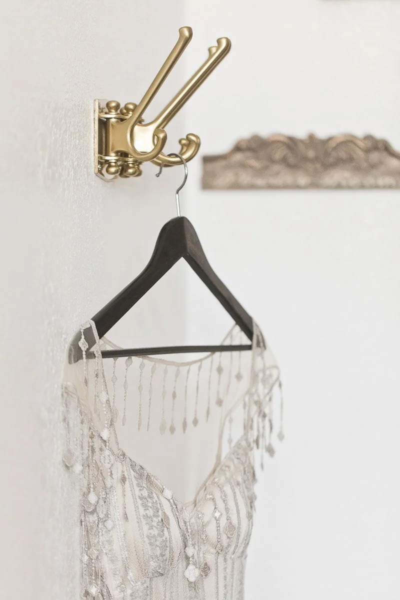 A shimmering white dress with intricate beading hangs on a hook against a white wall, exuding elegance. A decorative molding is blurred in the background.