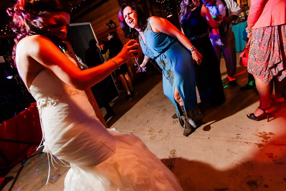 A joyful bride in a white gown and a woman in a blue dress enthusiastically dance on a lively, colorful floor during a wedding reception.