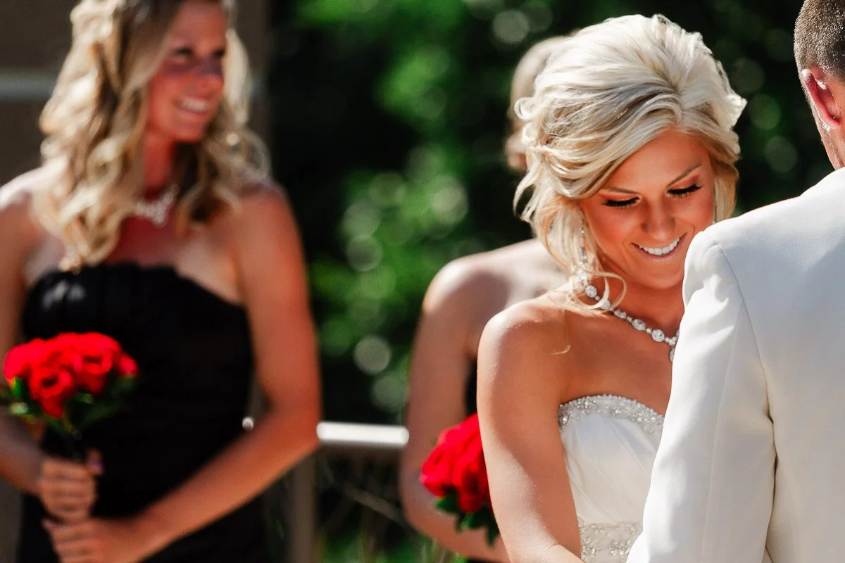 A bride in a white gown smiles during an outdoor wedding ceremony, holding hands with a man. Bridesmaids in black dresses with red bouquets look on.