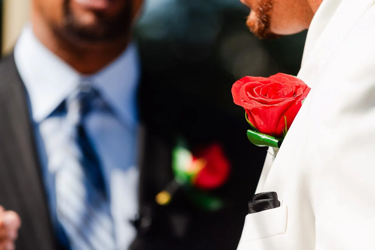 Close-up of two people in formal attire. One wears a white suit with a bright red rose boutonniere, and the other is in a dark suit with a tie, creating a festive and elegant tone.