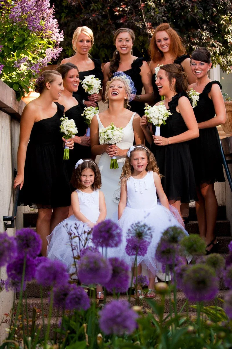 A bride laughing joyfully, surrounded by bridesmaids in black dresses holding white bouquets. Two young flower girls in white stand in front, with purple flowers in the foreground, creating a vibrant, celebratory scene.