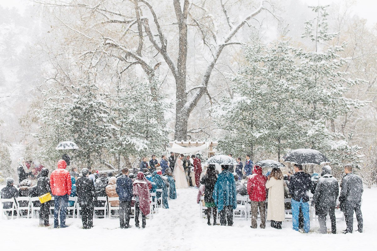 Snowy outdoor wedding ceremony with guests in coats and umbrellas, gathered around a couple under a tree. Peaceful, festive, and romantic atmosphere.