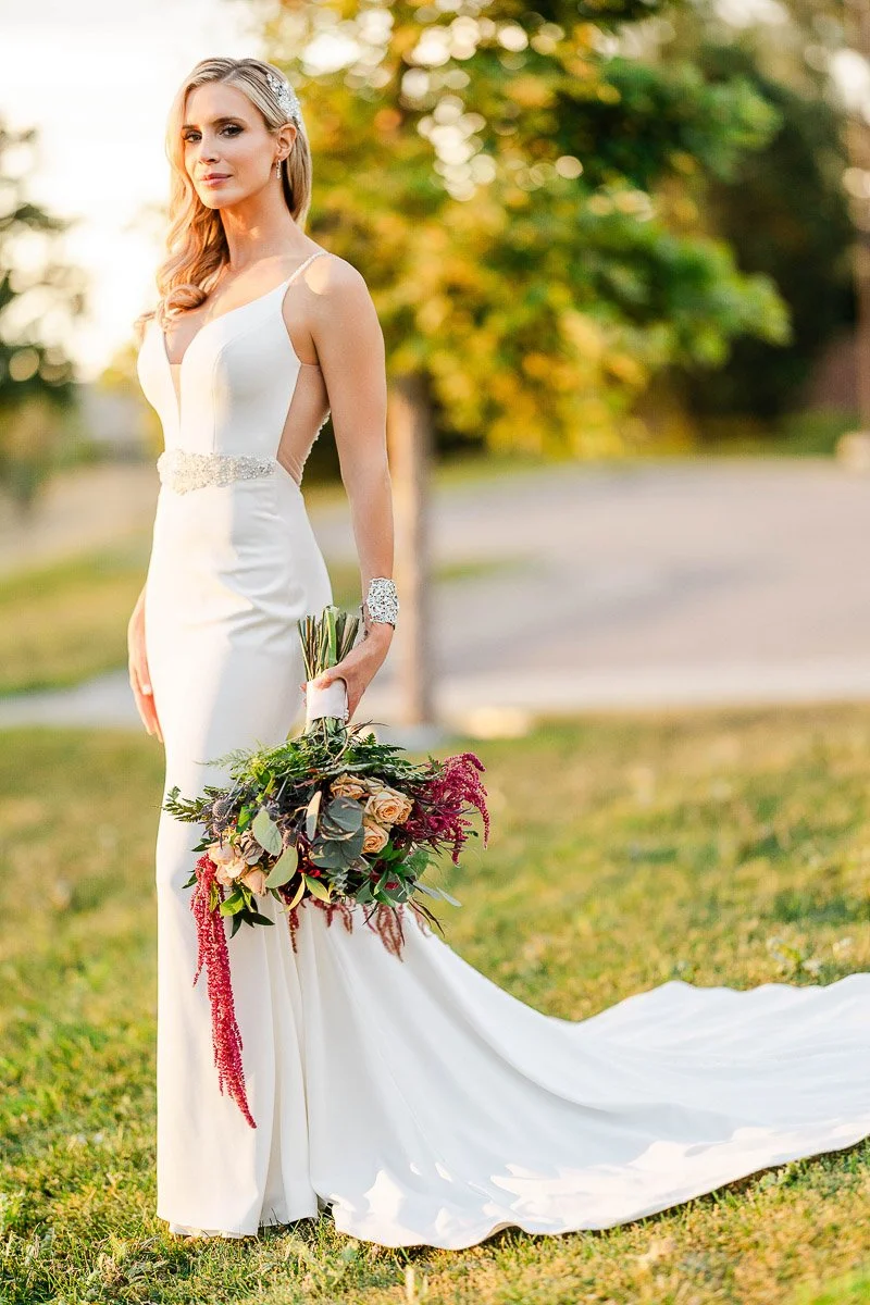 Bride in a sleek white gown with lace details and a long train stands on grass, holding a vibrant bouquet. Sunlight filters through trees, creating a serene atmosphere.