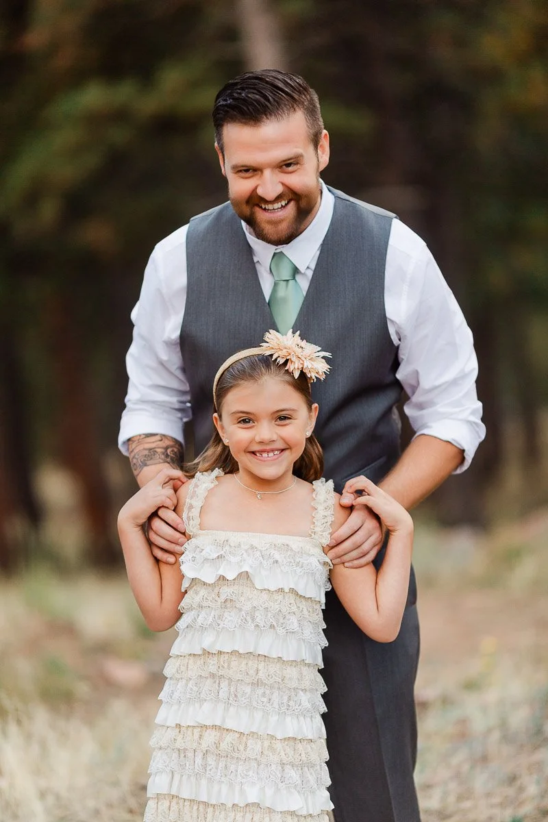 A smiling man in a gray vest and green tie stands behind a young girl in a lace dress with a flower headband, set against a blurred forest backdrop.