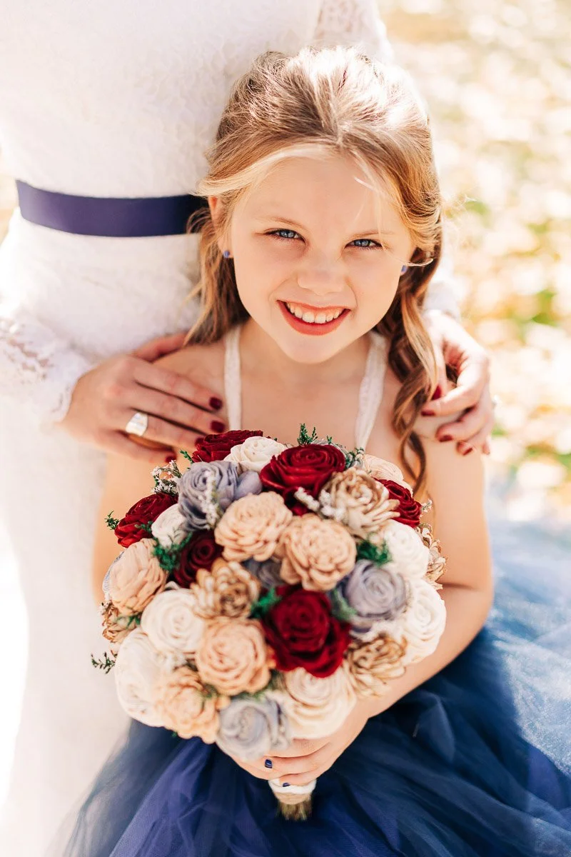 Smiling girl in a blue dress holds a colorful bouquet, standing with an adult in a white dress. The scene is joyful with soft, natural lighting.