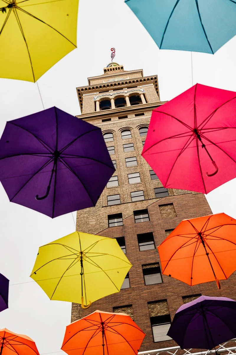 Colorful umbrellas, including yellow, pink, and blue, hang against a cloudy sky, framing a tall Denver Clock Tower, creating a whimsical scene.
