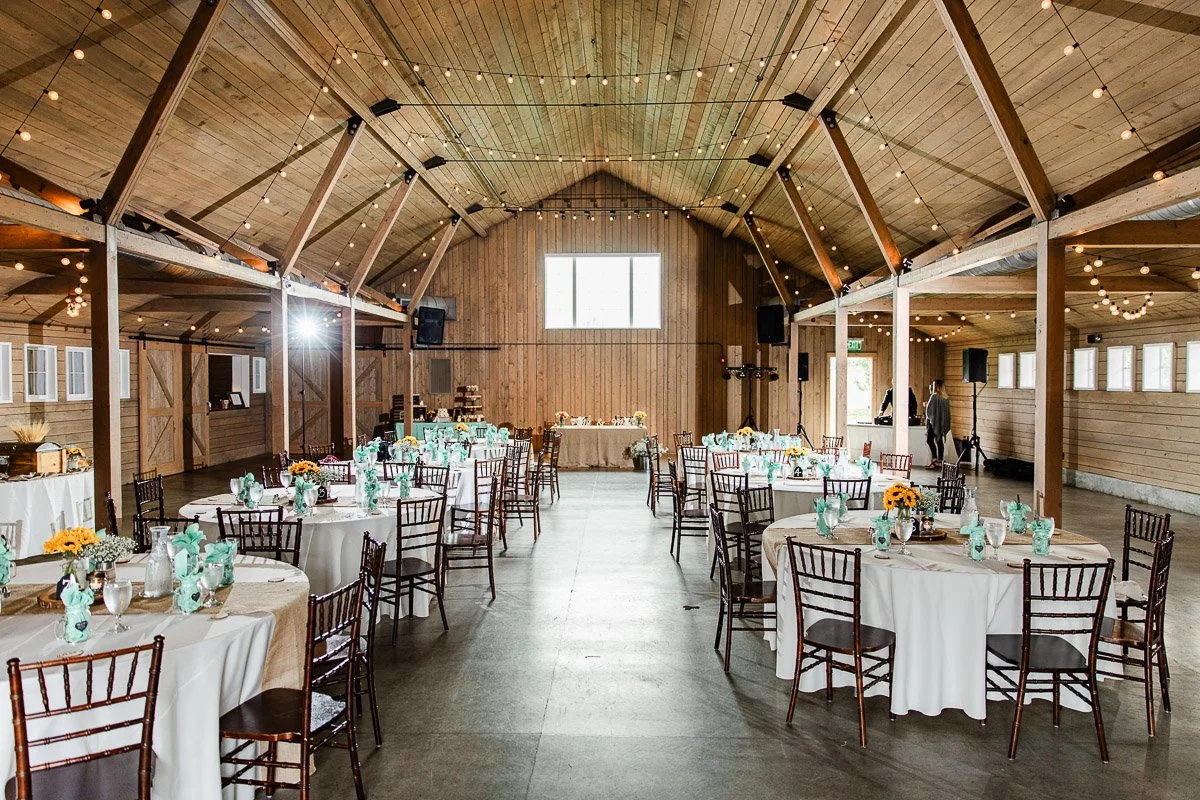 Rustic barn venue with wooden beams and string lights, set for an event. Round tables are decorated with teal napkins and sunflowers, creating a warm, inviting atmosphere.