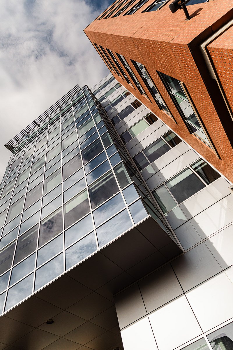 Upward view of a modern building with glass windows reflecting the cloudy sky, alongside a brick section. The perspective adds a dynamic, urban feel.