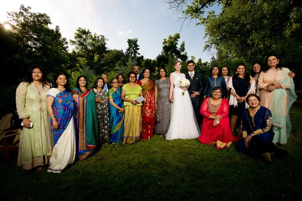 A diverse group stands on a lush lawn, featuring an array of colorful attire. A couple in wedding attire is centered, exuding joy against a backdrop of trees.