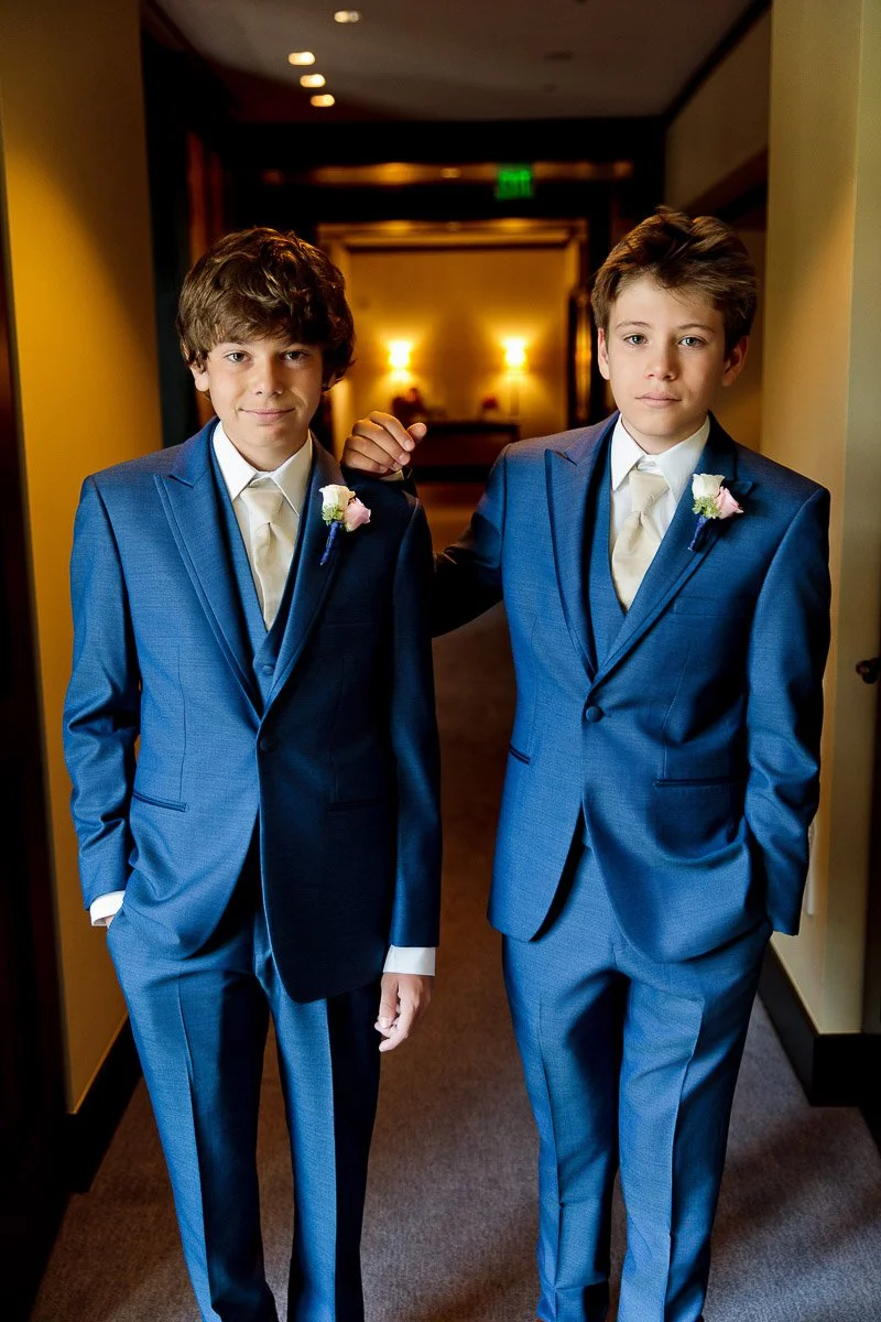 Two boys in blue suits with cream ties and floral boutonnieres stand in a warmly lit hallway. One boy rests his arm on the other's shoulder, smiling subtly.