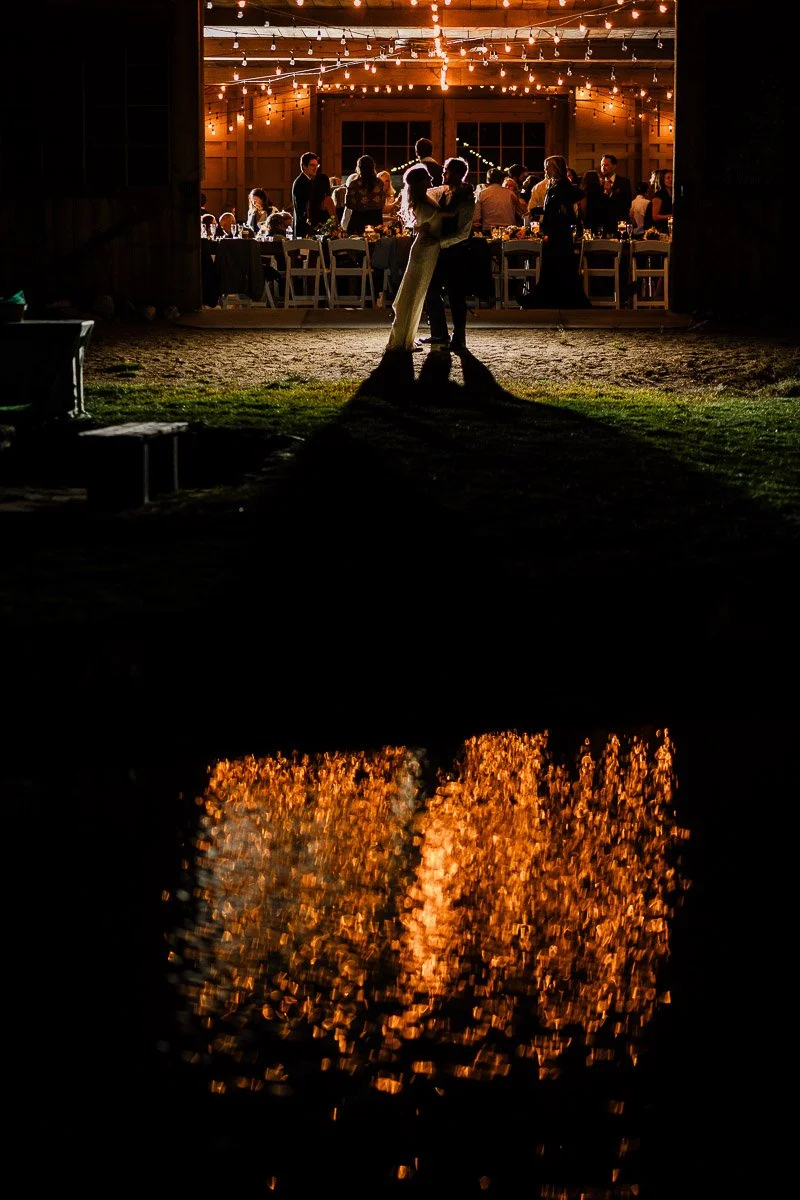 A silhouetted couple embraces under warm, glowing string lights inside a barn, with their reflection shimmering in dark water below, creating a romantic scene during a Strawberry Creek Ranch wedding in Granby, Colorado.