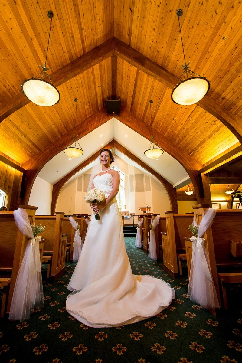 Bride in an elegant white gown holding a bouquet, standing in the aisle of a warmly lit church with wooden arched ceilings, conveying a joyful tone.