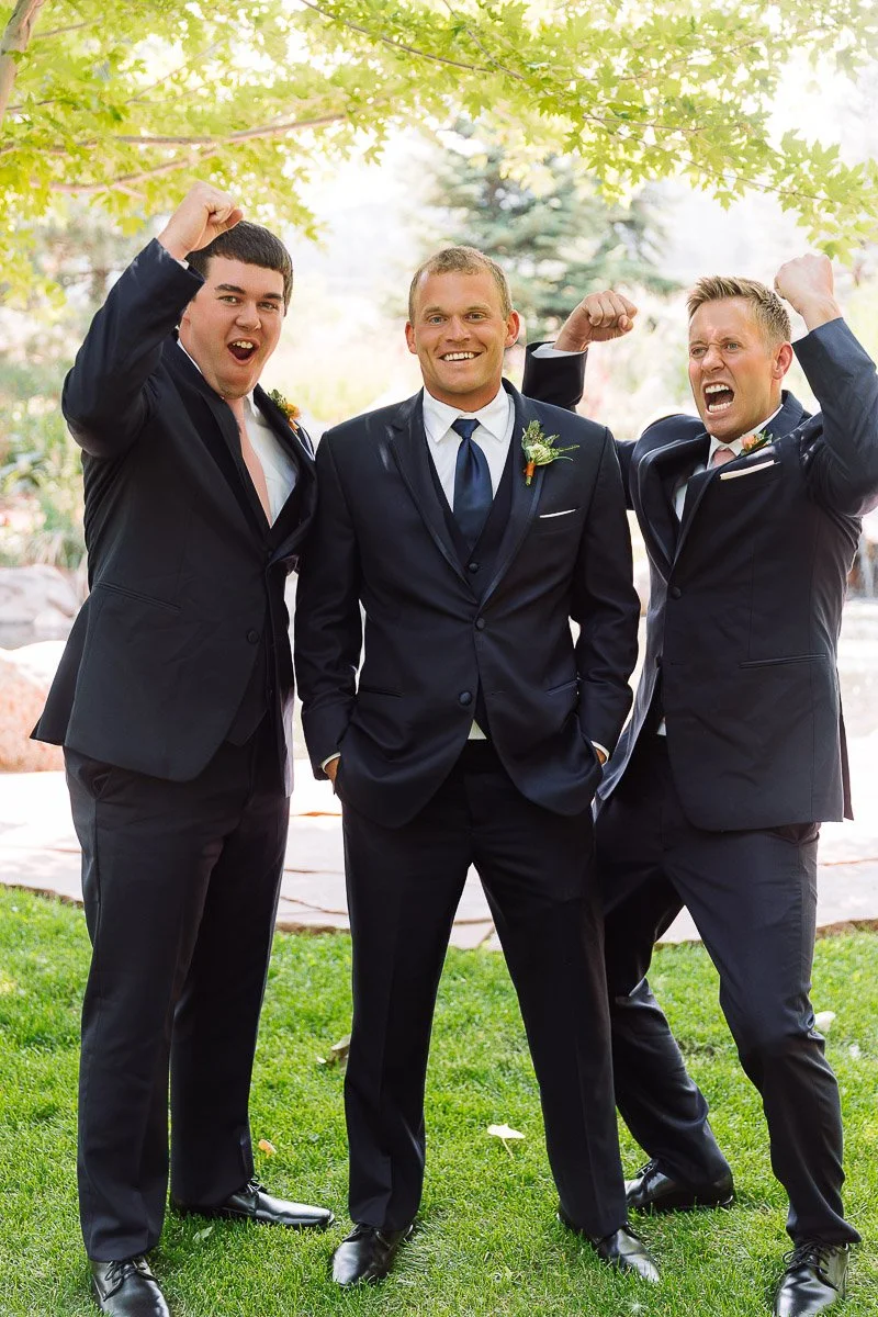 Three men in suits cheerfully pose outdoors on grass, surrounded by trees and sunlight. They display excitement and camaraderie, suggesting a celebratory occasion.