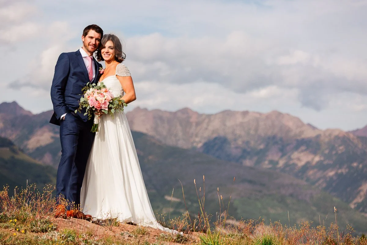 A joyful bride and groom stand on a grassy hill, with vivid mountains and a cloudy sky in the background. She holds a bouquet of pink flowers.