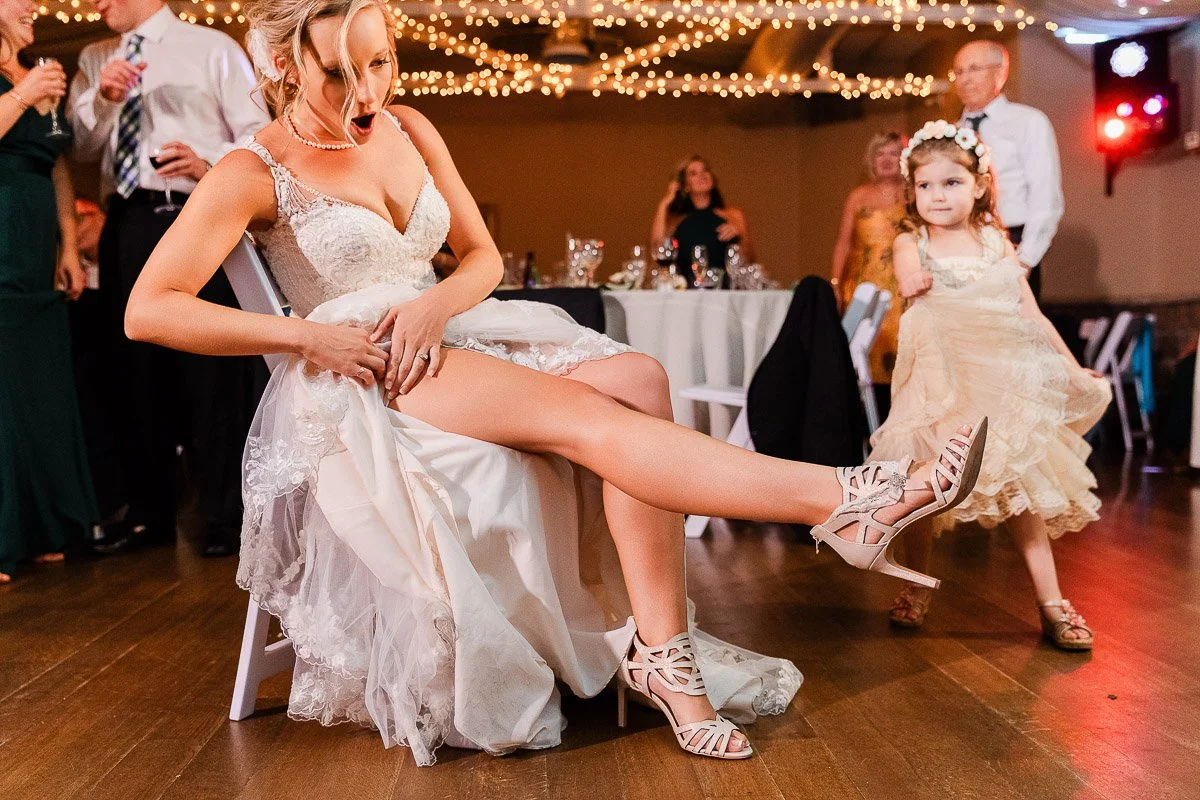Bride in a white lace dress sits on a chair during a lively reception, playfully revealing her garter. A flower girl in a beige dress runs by, amid guests and warm, glowing string lights.