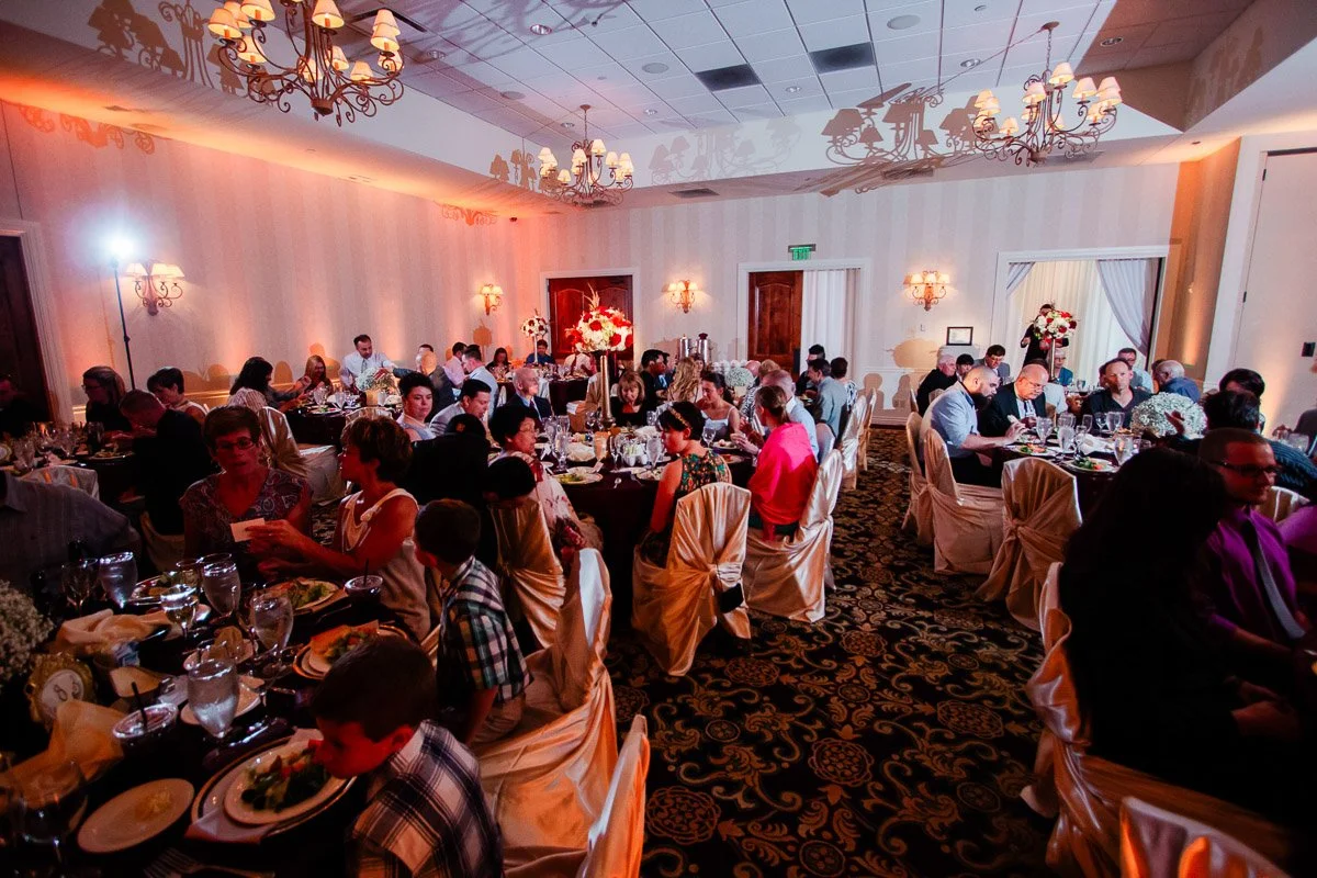 A warmly lit banquet room filled with guests seated at round tables. Elegant decor and floral centerpieces create a festive atmosphere, evoking celebration.