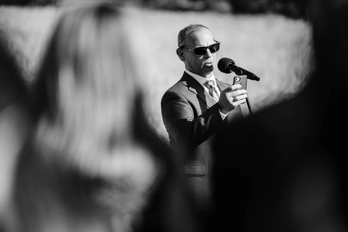 Man in a suit and sunglasses speaks into a microphone while looking at a phone, outdoors. Blurred foreground figures suggest a public event.