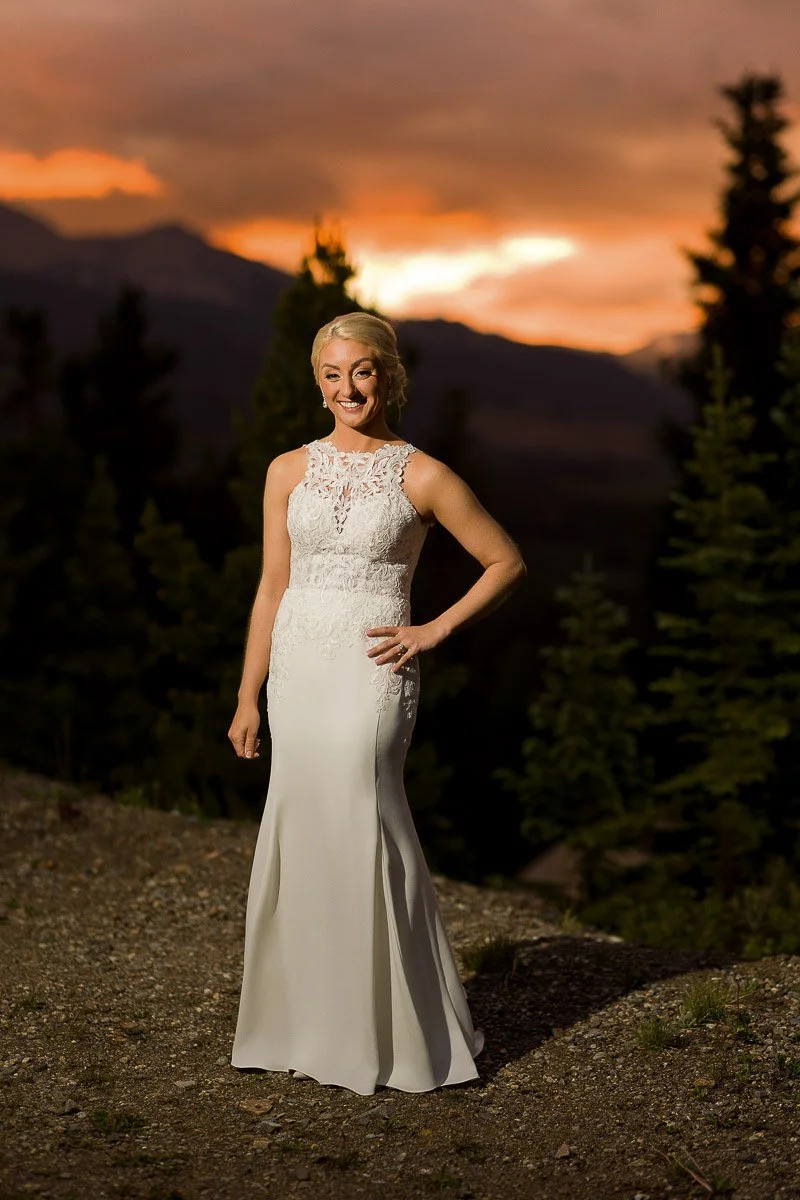 A woman in an elegant white lace wedding dress stands outdoors at sunset, smiling confidently. Dark trees silhouette against the vibrant orange sky.