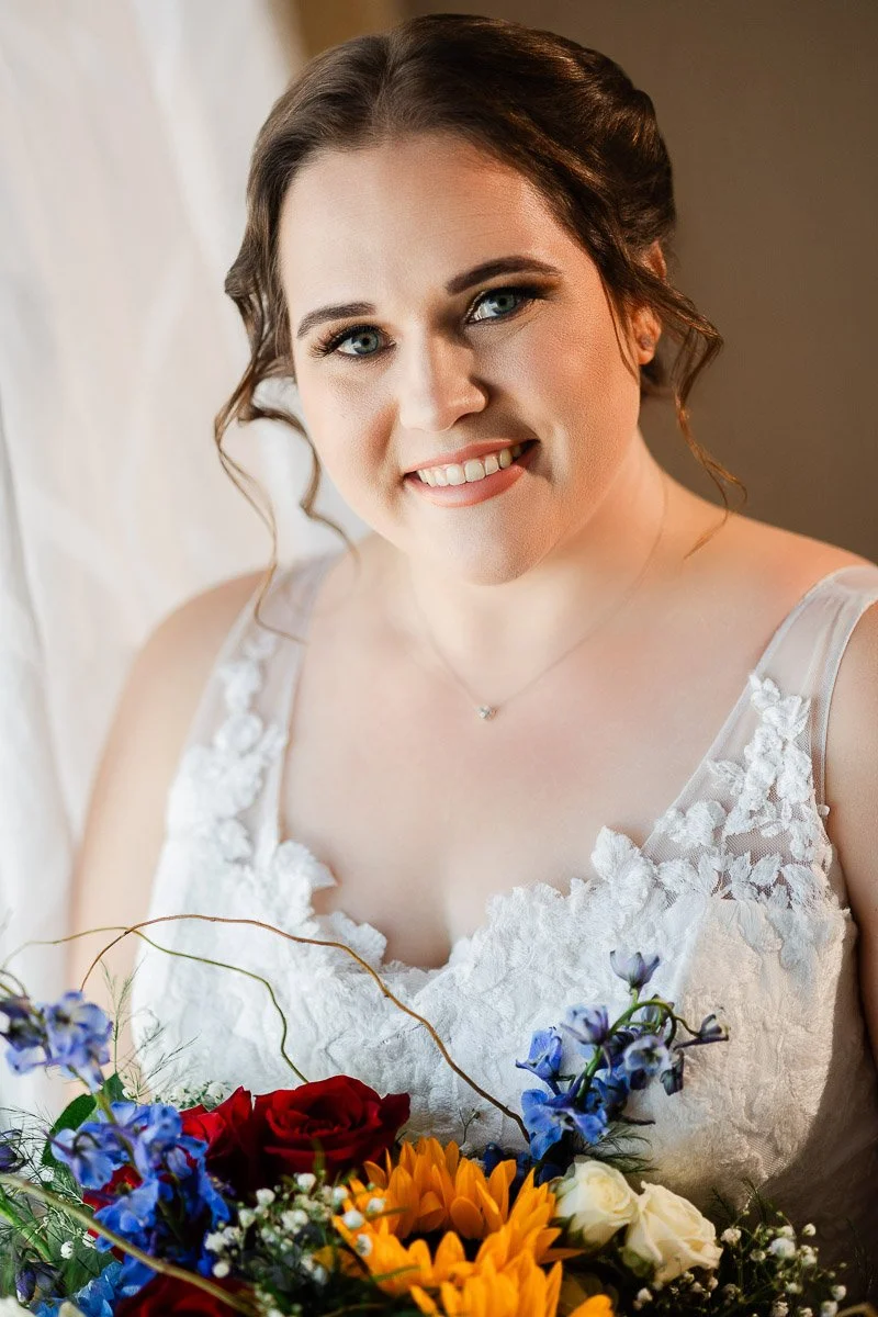 Smiling bride with an elegant updo and lace bridal gown holds a vibrant bouquet of sunflowers, red roses, and blue flowers, exuding joy and warmth.