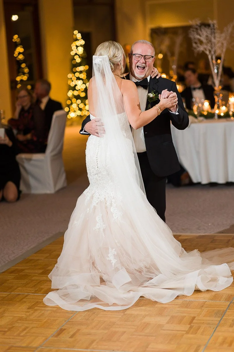 Bride and older man joyfully dancing at a wedding, with twinkling Christmas trees in the background and guests seated at tables, conveying warmth and celebration.