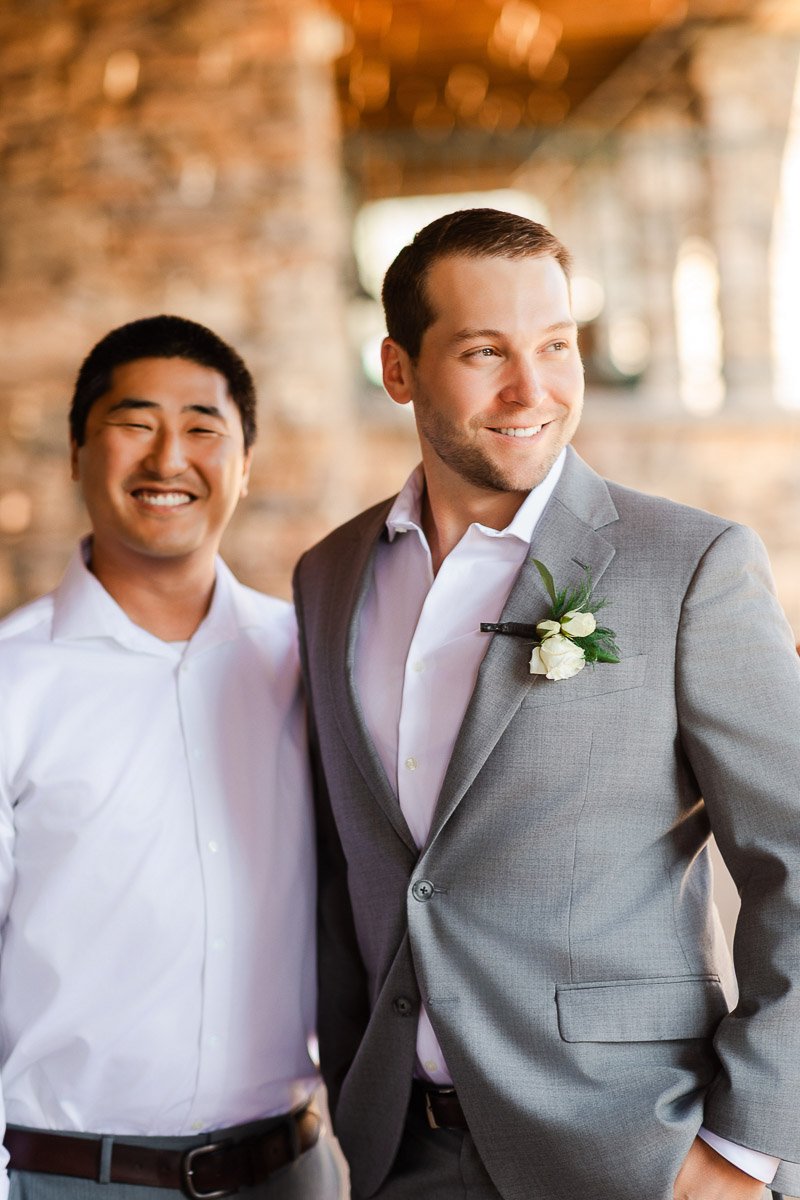 Two smiling men stand side by side. One in a gray suit with a boutonniere, and the other in a white shirt. Warm lighting creates a joyful, celebratory mood.