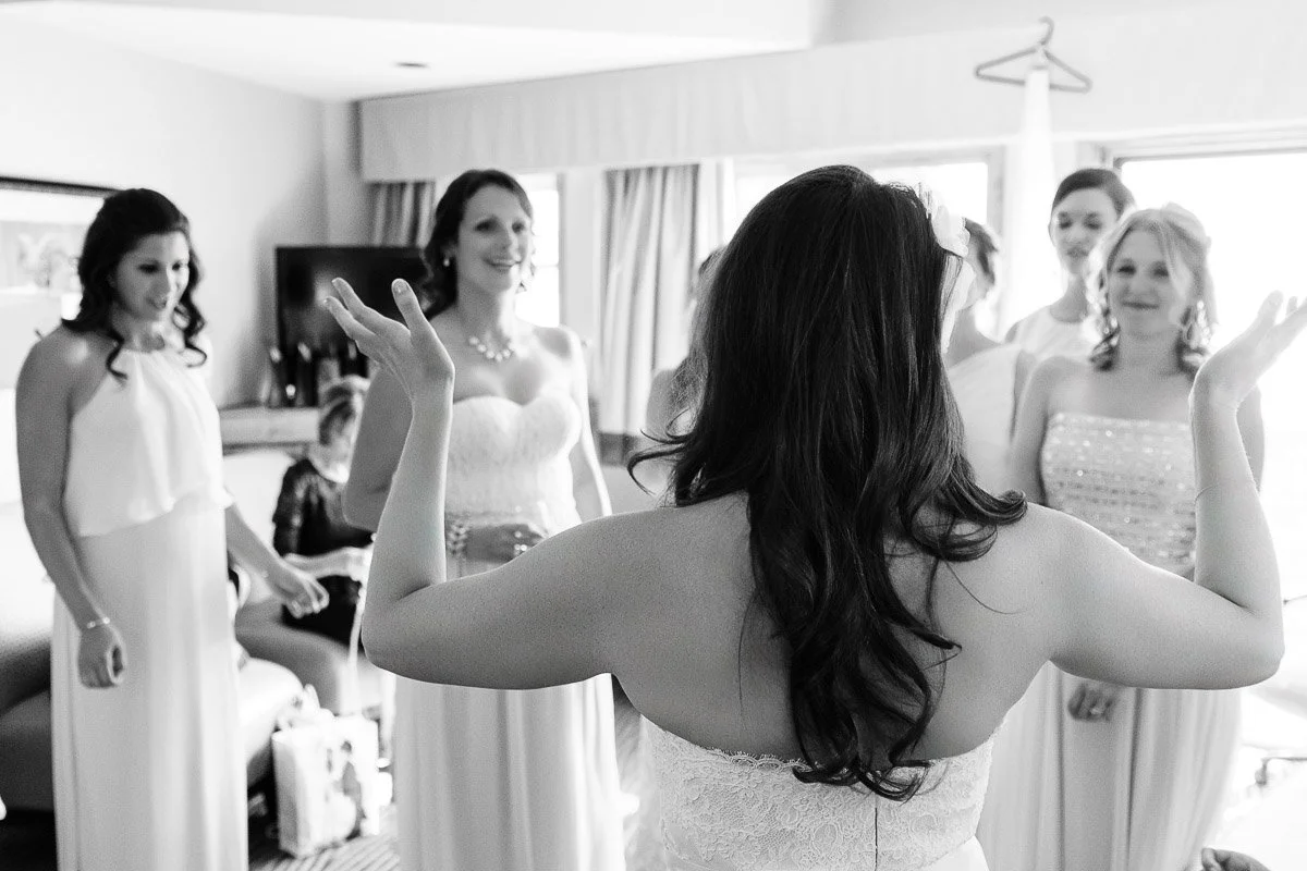 A bride with long hair, viewed from behind, joyfully raises her hands while talking to four bridesmaids in elegant dresses in a bright room.