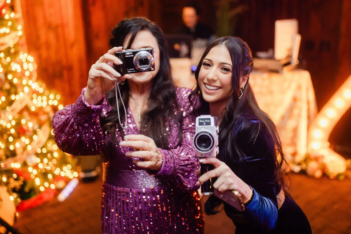 Two women smiling and holding cameras at a festive indoor event. One wears a sparkling purple dress, the other in black. A lit Christmas tree is in the background.