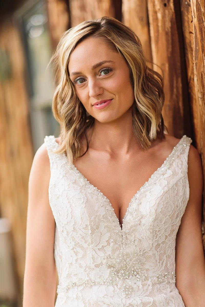 Smiling bride in lace dress with beaded detail poses against rustic wooden wall. Her wavy blonde hair complements the natural, joyful setting.