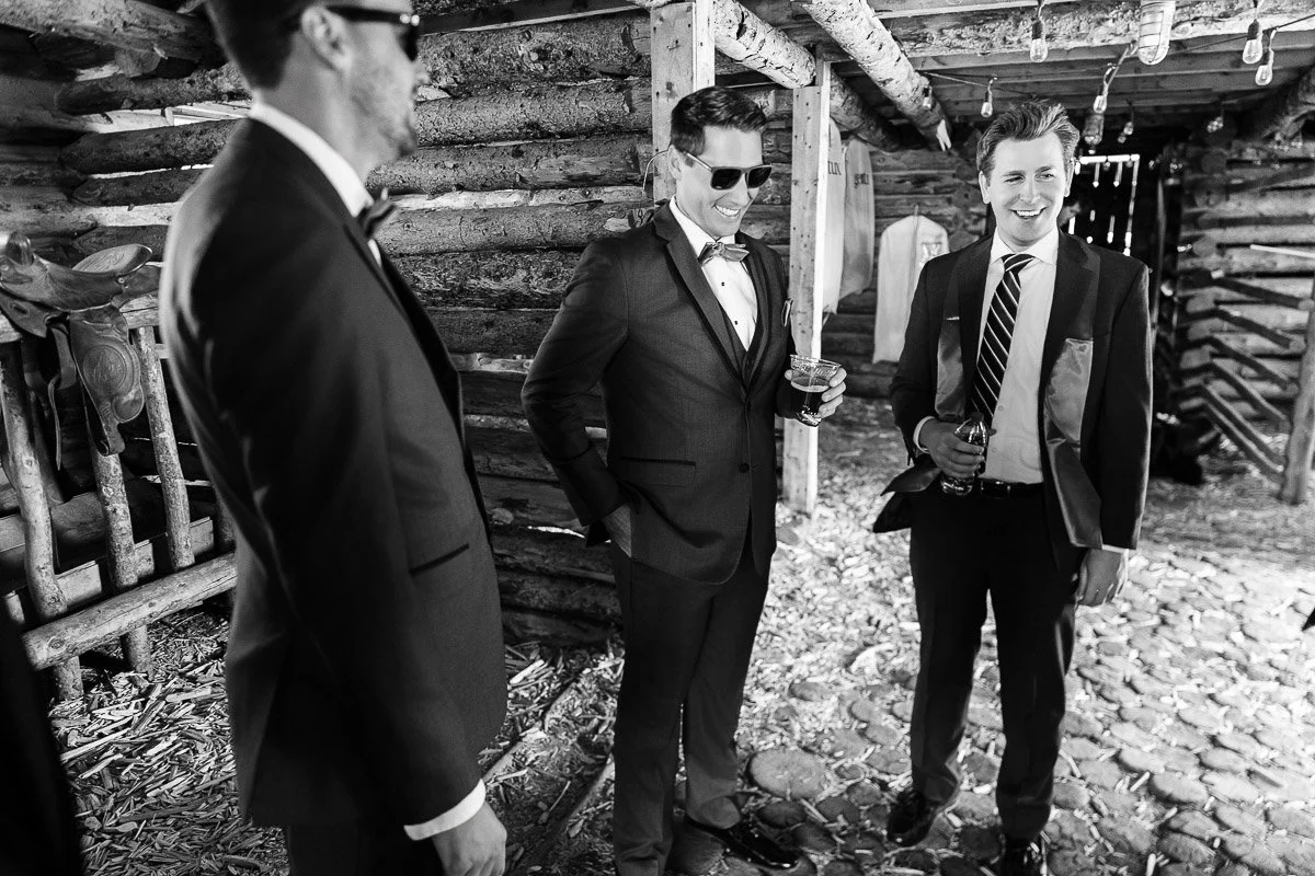 Three men in suits and sunglasses share a laugh in a rustic, log cabin setting. The mood is lighthearted and jovial, with drinks in hand.