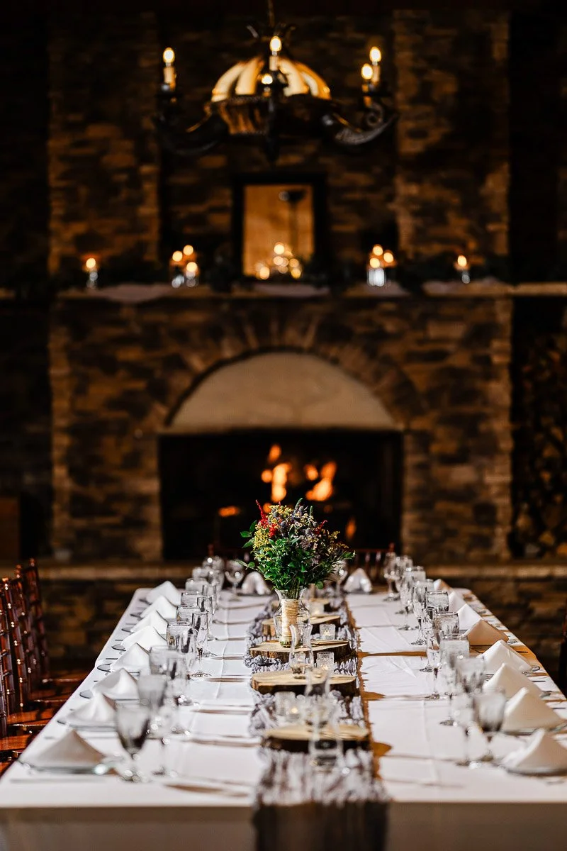 Elegant dining table set with white linens, glassware, and candles, centered with a floral arrangement. A warm, lit fireplace in the background.