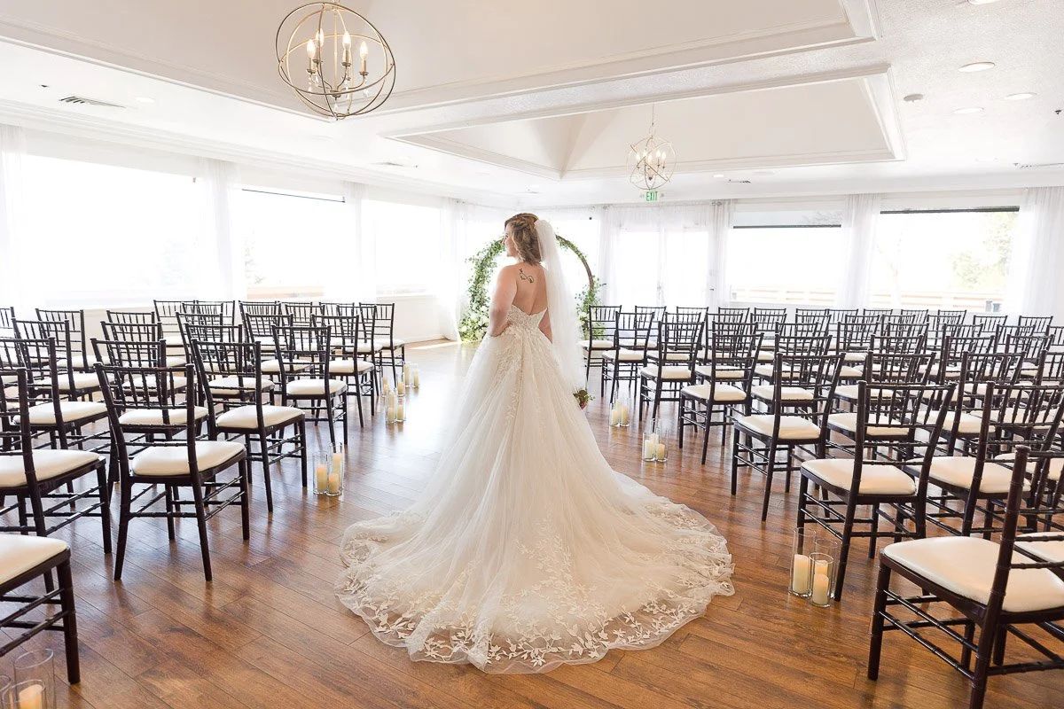 A bride in a flowing white gown stands in an empty, sunlit wedding venue. Rows of black chairs line the wooden floor, with candles placed alongside.