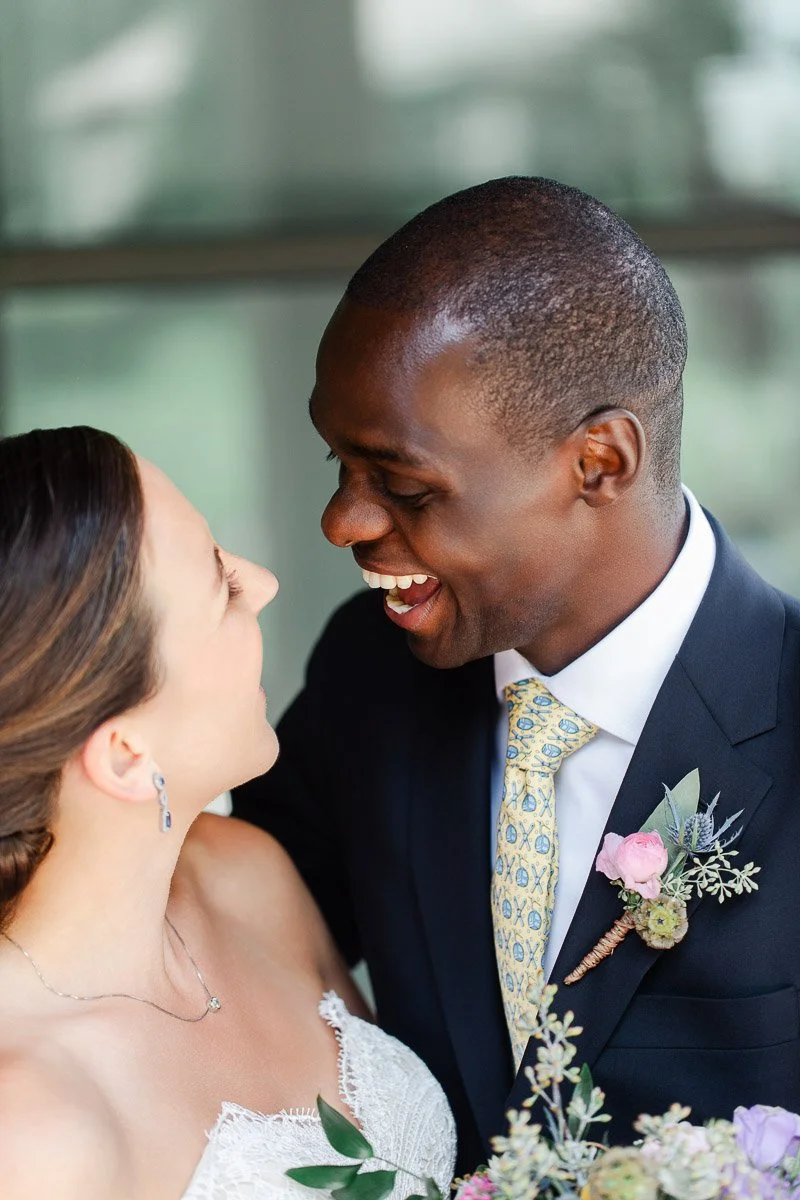 A joyful couple on their wedding day, face-to-face and smiling. The man wears a dark suit and yellow tie with a boutonniere. The woman is in a white lace dress.