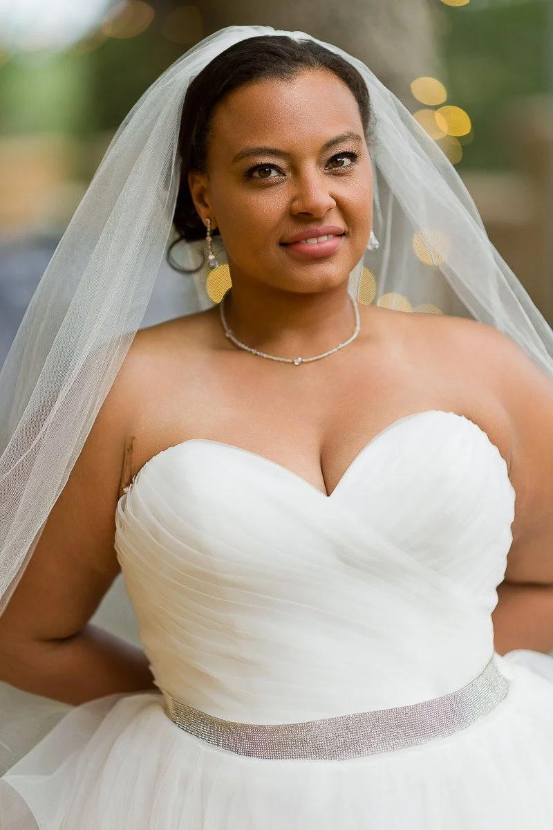 A bride in a white wedding dress stands confidently outdoors, wearing a veil and delicate jewelry. Soft lights twinkle in the blurred background, creating a serene, joyful atmosphere.