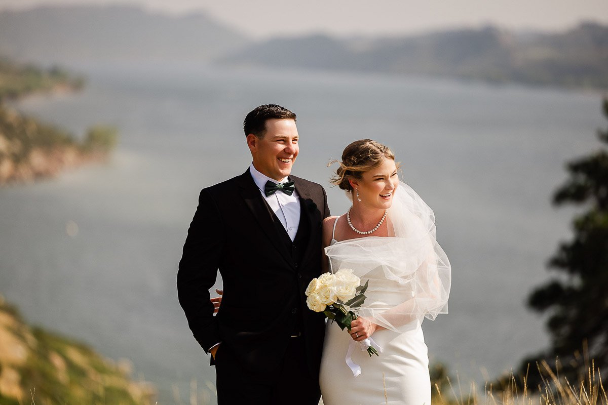 A bride and groom smile joyfully, standing on a grassy hill with a serene lake in the background. The bride holds a bouquet of white roses.