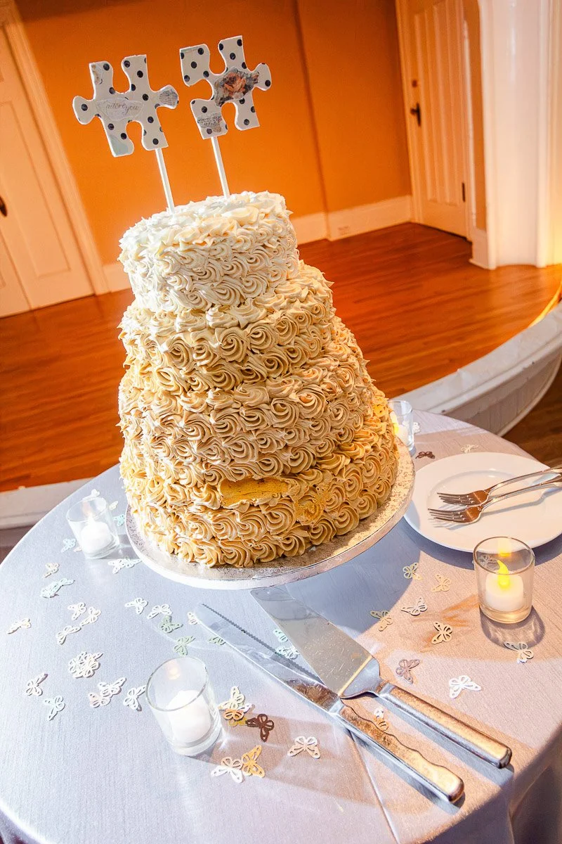 Three-tiered cake adorned with intricate swirls on a round table, topped with puzzle piece decorations. Candlelit ambiance and cutlery add elegance.