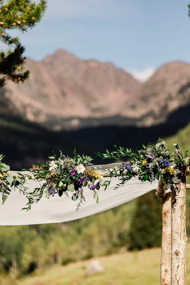 Wedding arch with wildflowers and greenery against a backdrop of majestic mountains under a clear blue sky, creating a serene, romantic setting.