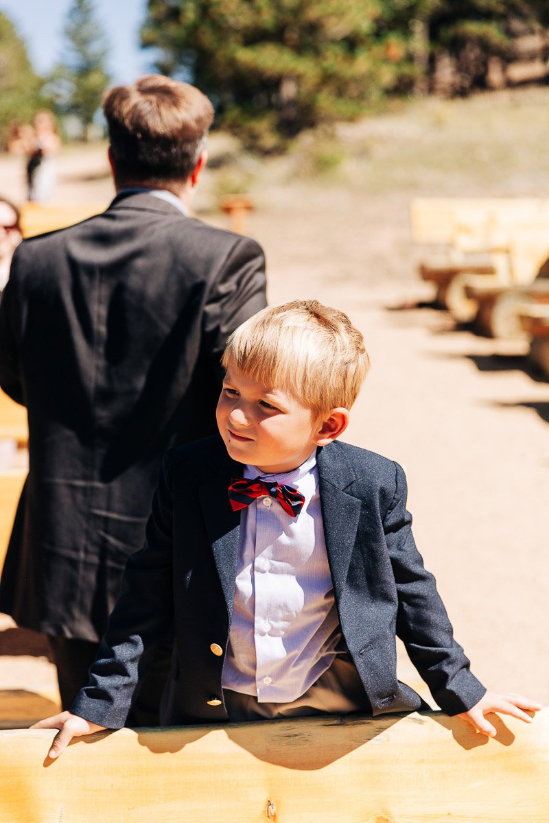 A young boy in a navy suit and red bow tie stands outdoors, leaning on a wooden railing. Sunlit trees and a clear sky create a warm, cheerful atmosphere.