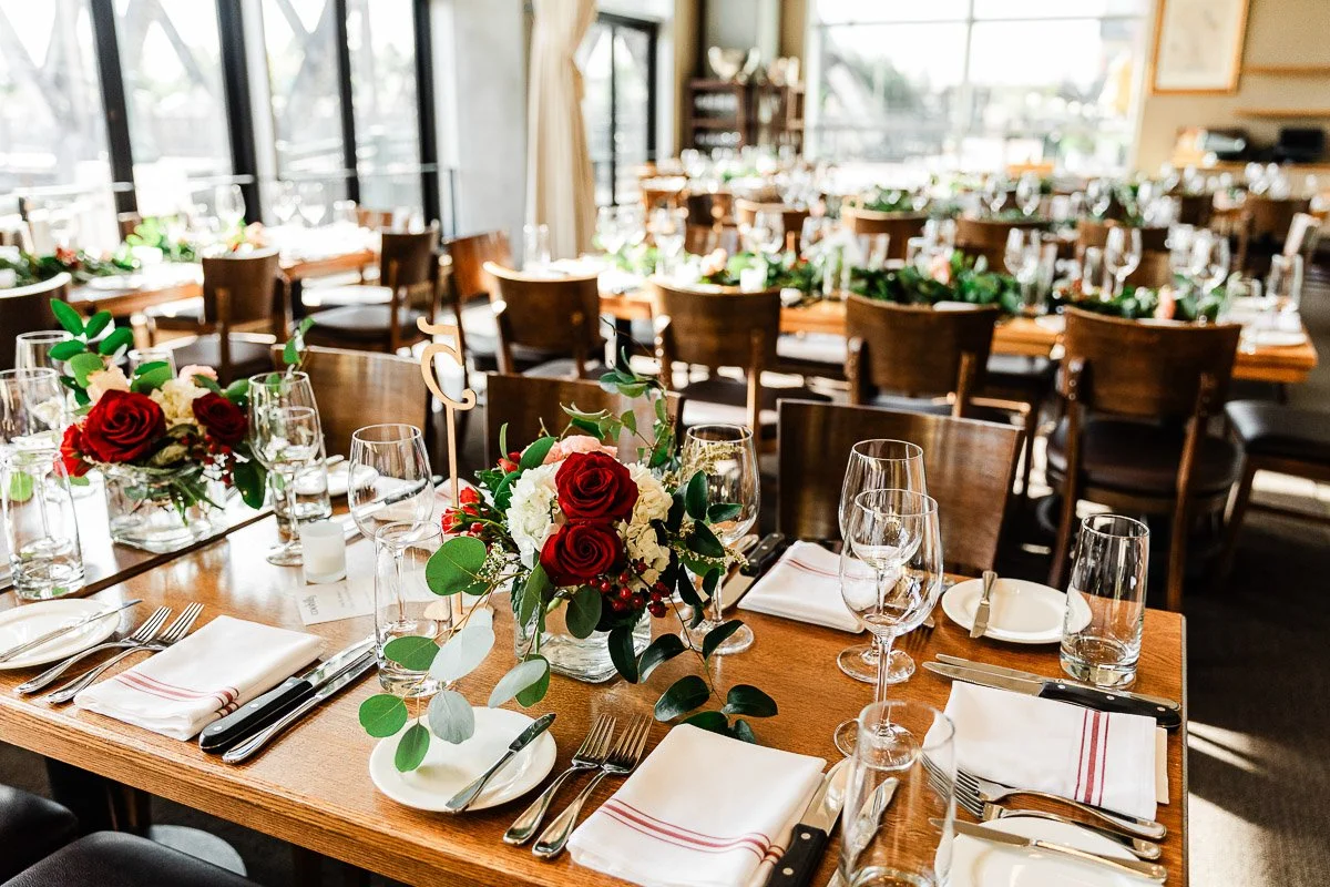 Elegant dining setup with neatly arranged wooden tables featuring red rose centerpieces, glassware, white napkins, and sunlight streaming through large windows.