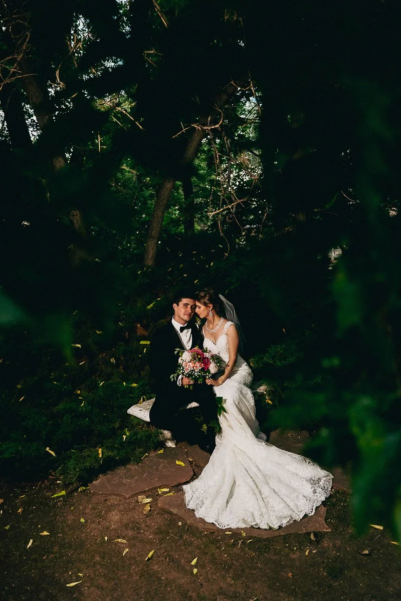 Bride in a white gown and groom in a black suit sitting on a bench in a lush garden. They share an intimate moment, surrounded by vibrant greenery.
