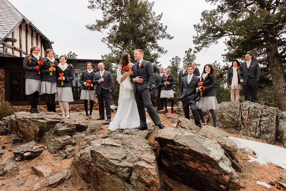 Wedding party on rocky terrain with trees and a rustic building. Bride and groom share a moment at a Boettcher Mansion wedding, surrounded by bridesmaids and groomsmen holding flowers. Celebratory and joyful.