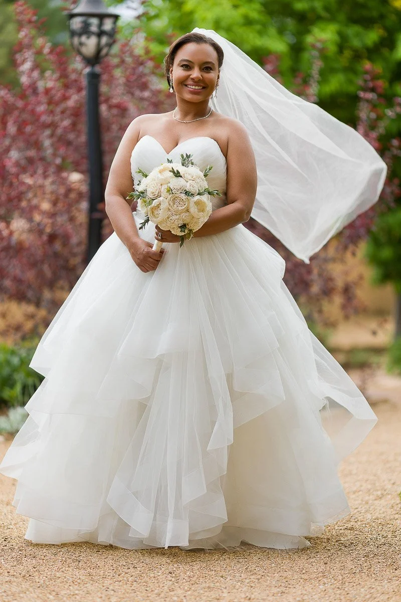 A joyful bride in a flowing white gown and veil holds a bouquet of white roses, standing on a garden path with lush green and burgundy foliage.