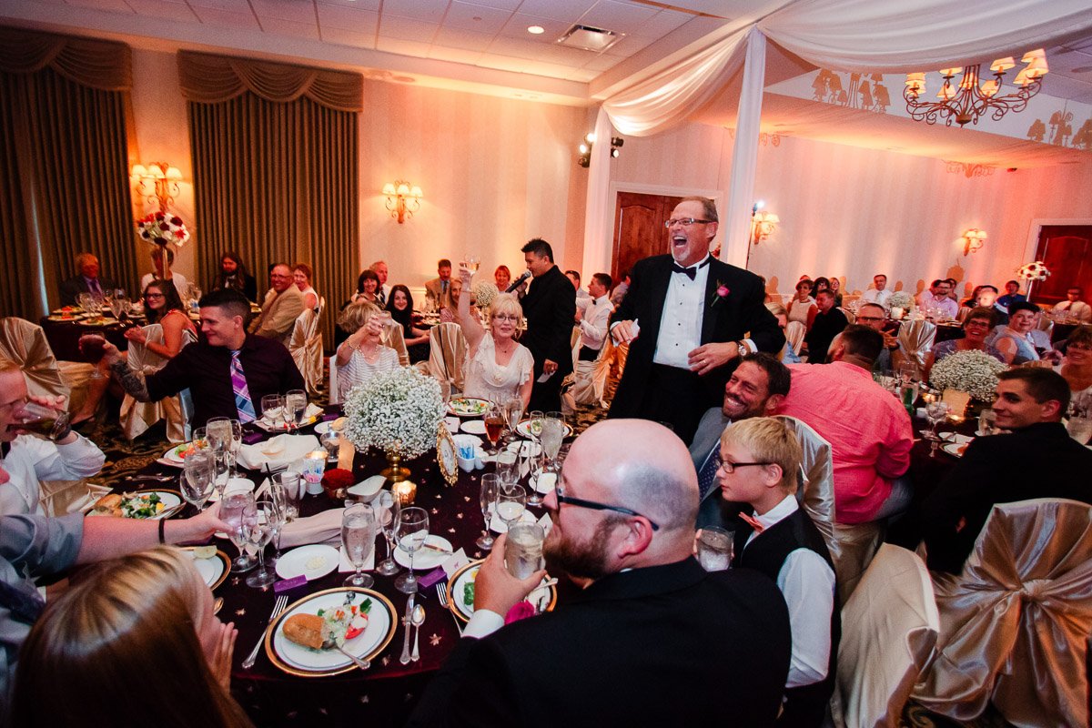 A joyful wedding reception with guests seated at round tables adorned with white flowers. A man in a tuxedo stands laughing, creating a lively atmosphere.