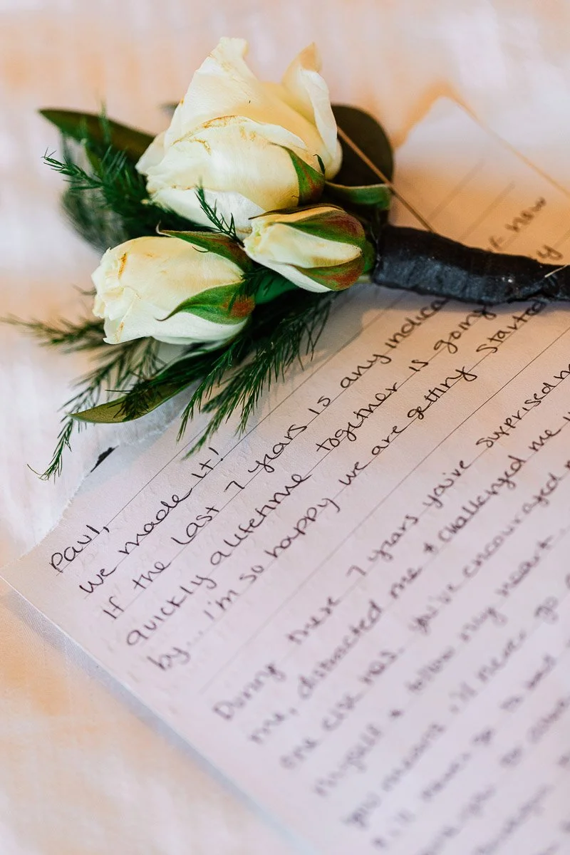 A handwritten letter lies on a white fabric surface, adorned with white roses and greenery. The tone is romantic and intimate.