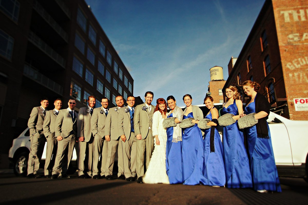 A smiling wedding party in formal attire poses on a sunny street. Groomsmen wear gray suits, bridesmaids in royal blue dresses, creating a joyful atmosphere.