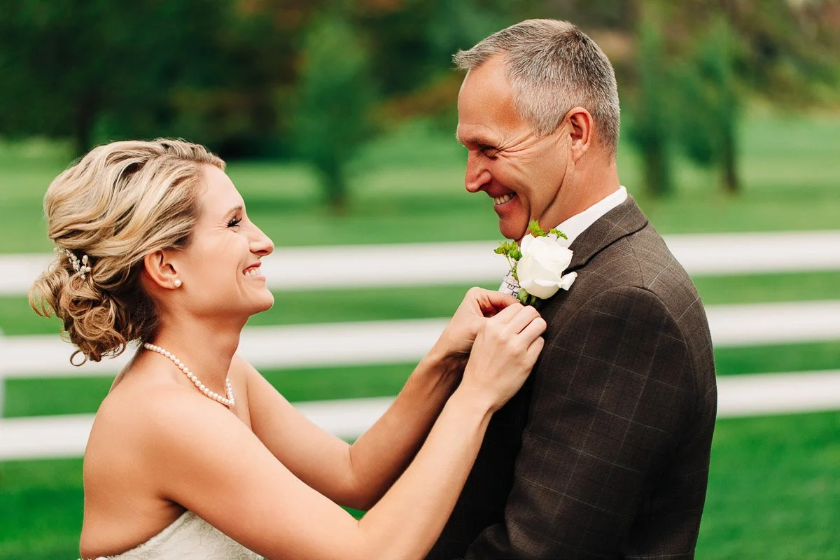 A joyful bride adjusts the groom's boutonniere on a sunny day. They are smiling warmly at each other in an outdoor setting with green grass and a white fence.