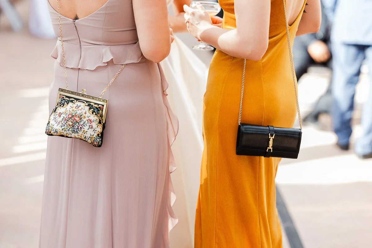 Two women at an event, one in a beige dress with a floral-patterned purse, the other in an orange dress with a black purse, holding drinks. The scene conveys elegance.