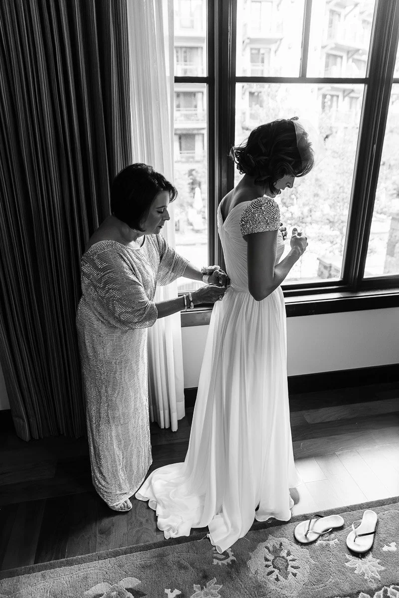 A woman in a gown is getting assisted with her dress by another woman near a large window. The scene is calm and intimate, with soft natural light.